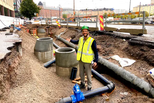 Endrit Kola in a hard hat and high-visibility vest, holding a notebook, standing in a street trench in central Helsinki with new manholes, water pipes and a valve.