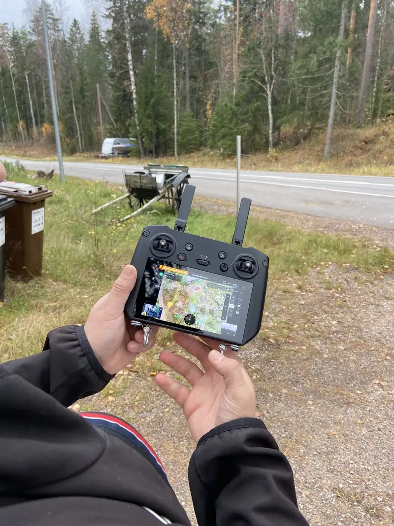 Hands holding a drone controller with a map on its screen, beside a forest road.