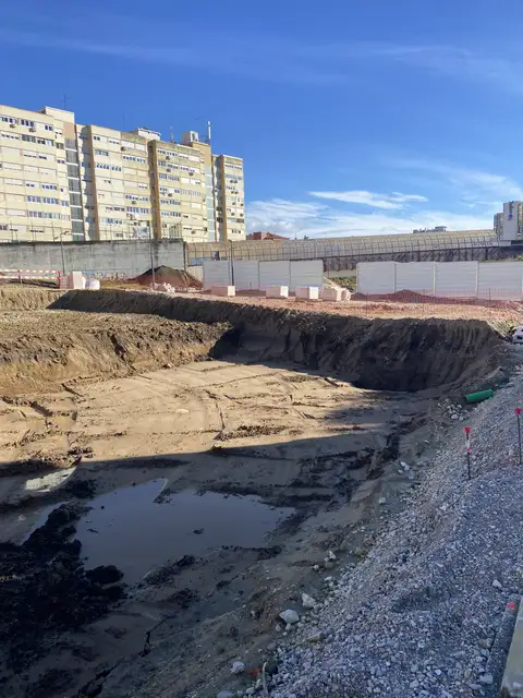 Wide open excavation with standing water, beside apartment blocks.