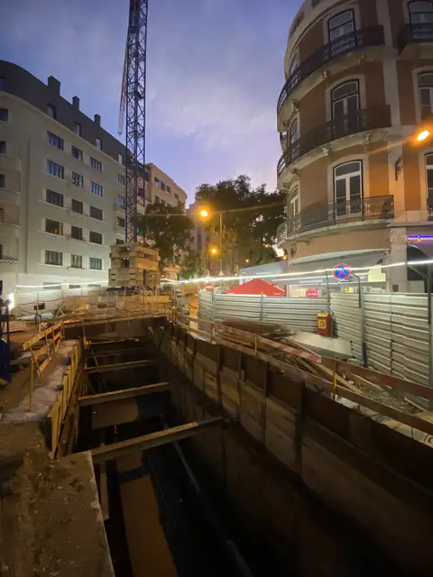 Shored excavation along a city street at night, with a crane behind.