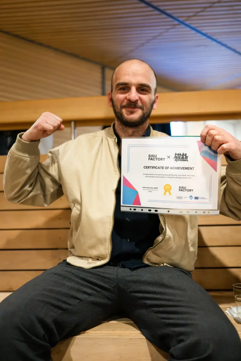 Endrit Kola seated on a sauna bench, holding the Sisu Factory certificate for winning Sauna Pitching.