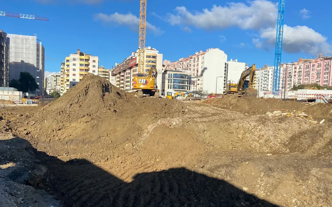 Large urban earthworks site with spoil heaps, two excavators and two tower cranes under a blue sky, apartment blocks behind.