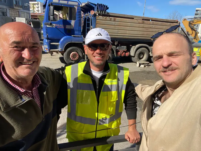 Three people smiling on a construction site, one in a high-visibility vest marked Inxhinier (engineer), with a dump truck and an excavator behind them.