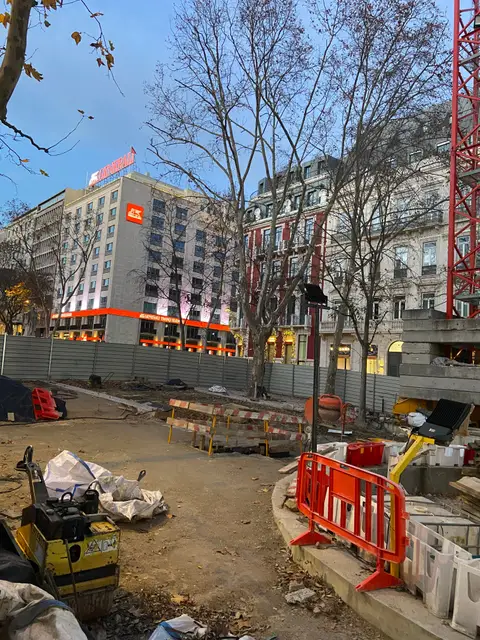 Wide view of a city construction site at dusk, with barriers, a compactor and a hotel behind the site fence.